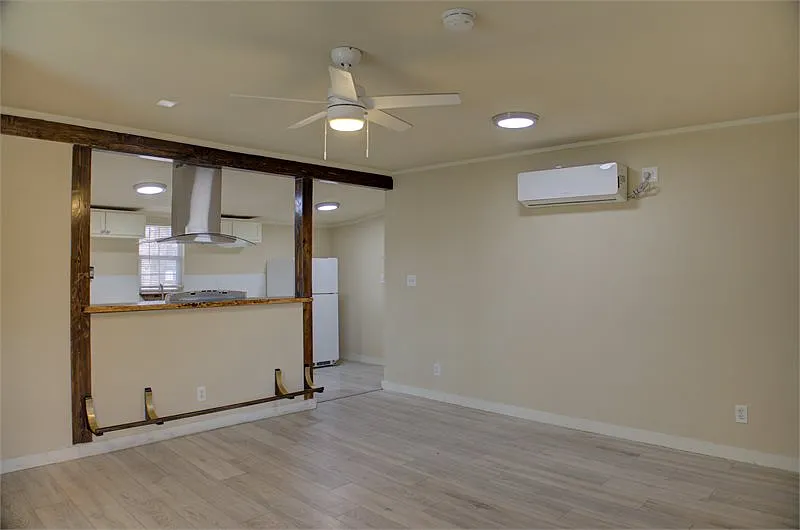 Empty living area with light wood floors, ceiling fan, and a wood-framed kitchen pass-through.