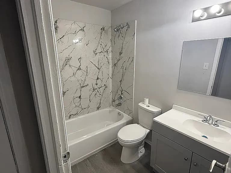 Clean bathroom with marble-patterned shower tiles, white bathtub, toilet, and a vanity with mirror.