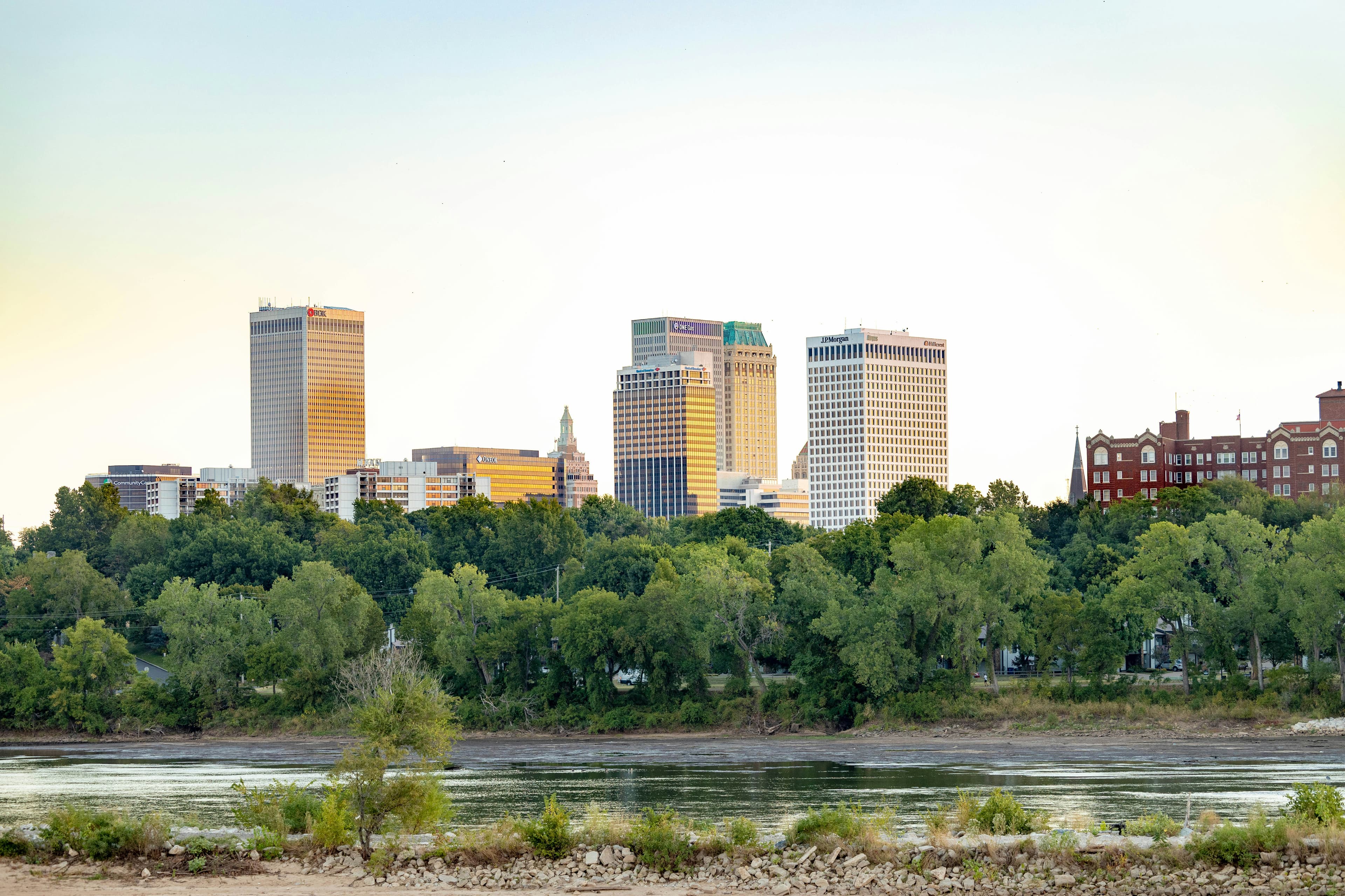 Tulsa city skyline with skyscrapers rising above lush green trees along a wide river.